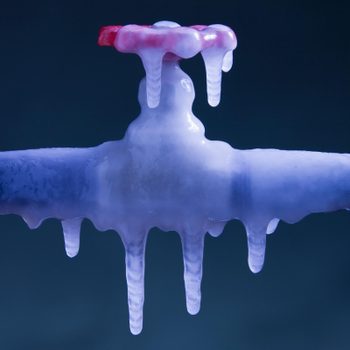 Frozen pipe with icicles hanging, surrounding the red valve. Dark background emphasizes the icy texture and cold environment.