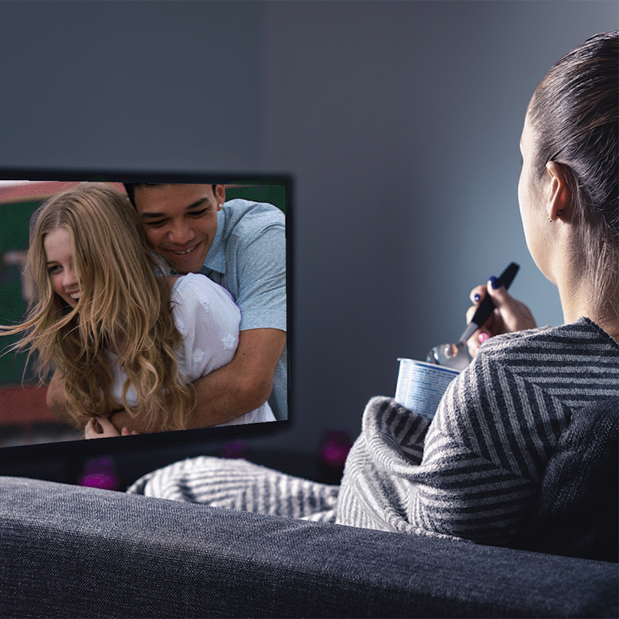 Person eats from a tub while watching TV, featuring a man hugging a woman, in a dimly lit living room.