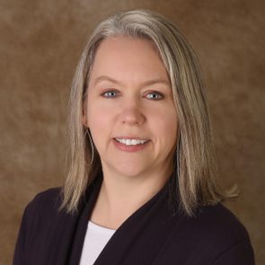 Person smiling against a brown textured background, wearing a dark cardigan and white shirt, with medium-length gray hair framing the face.
