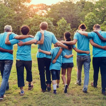 People in blue shirts walk with arms linked in a grassy park at sunset.