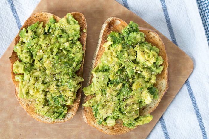 Over head avocado toast on slices of toasted French loaf bread topped with ground lemon pepper and sprinkles of kosher salt on wood cutting board on top blue and white striped linen cloth