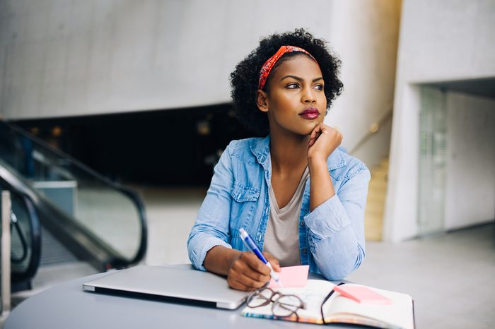Focused young African female entrepreneur deep in thought while working at a table in a modern office building lobby