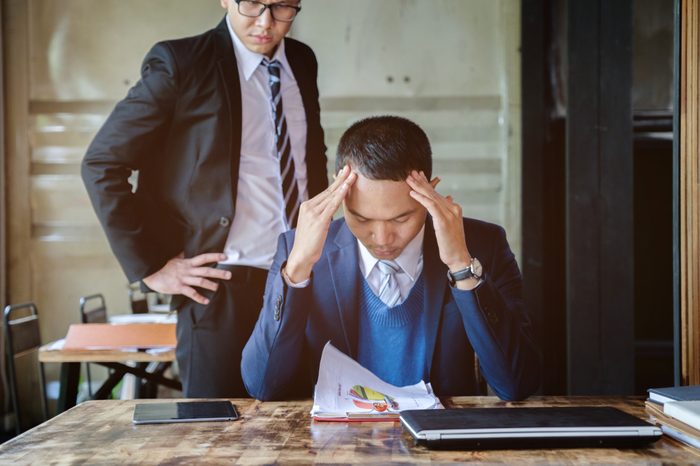 stressed asian business man working with laptop on desk, boss/manager do angry face complaining standing behind employee, business co-working unhappy teamwork concept
