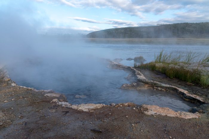 Steamy river water and muddy shoreline with green grass under blue sky with white fluffy clouds.