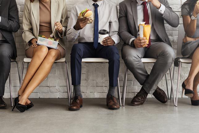 Four seated people eat food, including a sandwich, salad, and baguette, while dressed in business attire against a white brick wall backdrop.