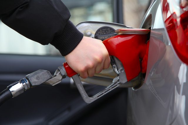 Hand grips red fuel nozzle, refueling silver car, with caution text visible on pump.