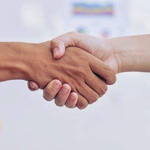 Two hands shaking, signaling agreement, with a blurred background containing colorful sticky notes and charts.