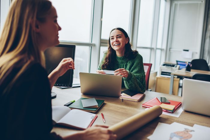 Smiling young women having fun during cooperation at desktop in office , happy female colleagues laughing at joke during working process using modern technology for collaborating on common project