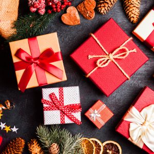 Gift boxes are arranged with ribbons on a dark surface, surrounded by festive items like pinecones, candy canes, dried oranges, and evergreen branches.