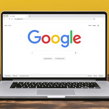A laptop displays Google's homepage on a wooden table, surrounded by a green plant and a yellow mug, against a yellow wall.