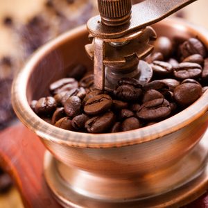 Coffee beans fill a manual grinder in close-up, surrounded by scattered beans on a wooden surface.