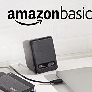 Speakers rest on a desk, connected to a device. The setting includes a laptop and headphone in a bright, minimalist office space. Text reads 