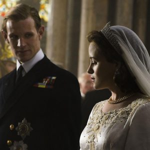 A woman in a wedding dress stands beside a man in a uniform, inside a church with stained glass windows softly illuminated.