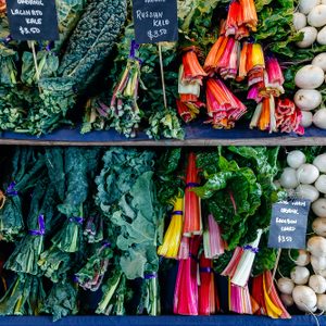 Vegetables displayed on shelves with labeled signs showing prices, featuring beets, kale, chard, and radishes in a market setting.