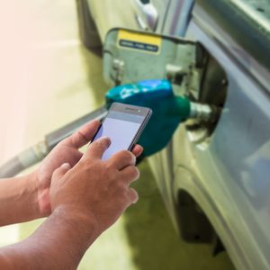Hands hold a smartphone beside a car being refueled with a blue gas nozzle at a gas station.