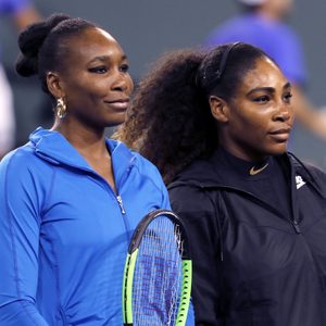 Two women stand together, one holding a tennis racket, on a sports court, wearing blue and black athletic jackets.