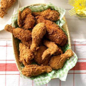 Fried chicken fills a woven basket, lined with green checkered paper, beside a table with red checkered cloth and yellow forks.