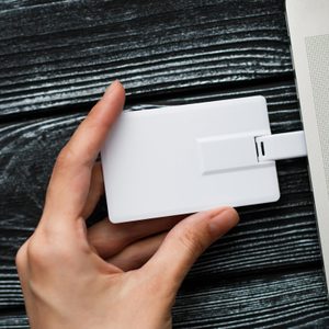 A hand holds a white USB card, plugged into a silver laptop, on a dark wooden surface.