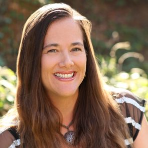 Smiling woman stands outdoors, wearing a necklace, with blurred greenery in the background.