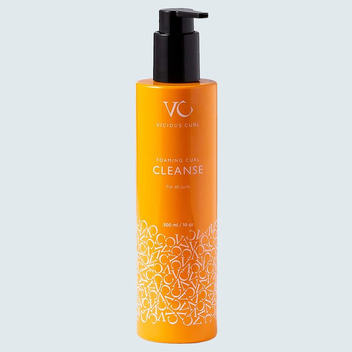Bottle with pump dispenses foaming curl cleanser, orange with white text and patterns, against a light gray background.