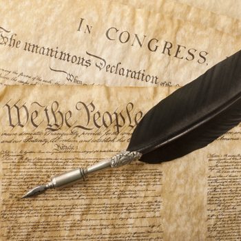 Quill pen rests on aged documents, including "We the People" text, suggesting historic American government papers.