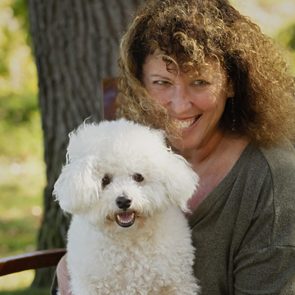 A fluffy white dog sits cheerfully on a woman's lap; they are outdoors near a tree with blurred greenery in the background.