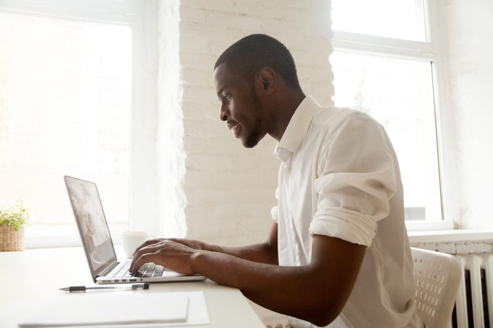 African businessman working on laptop sitting at home office desk, happy black intern employee looking at computer screen typing or browsing web, using software application for business and education