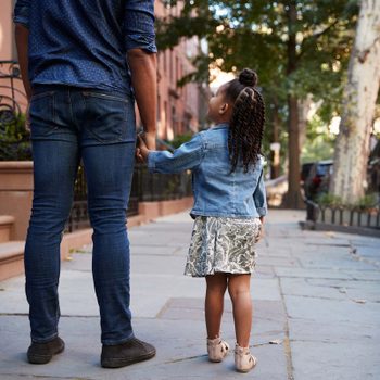 Adult and child walk hand in hand down a tree-lined sidewalk, bordered by parked cars and brownstone buildings.