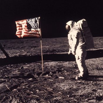 Astronaut stands beside raised American flag on the moon's surface, with lunar footprints and a spacecraft leg visible, under a dark sky.