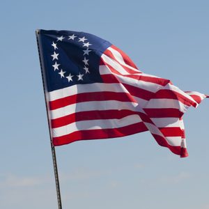 A flag with thirteen stars in a circular pattern waves against a clear blue sky.