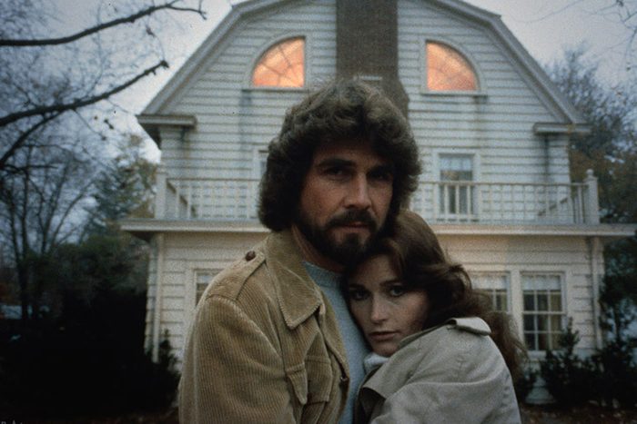 A couple stands embracing in front of a large white house with distinct upper windows, surrounded by trees on an overcast day.