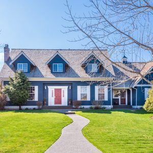 A blue house stands on a lush lawn with a winding path, surrounded by trees and shrubs under a clear, blue sky.