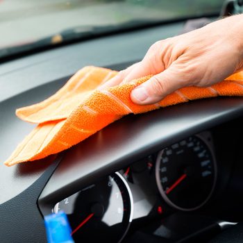 Hand wipes car dashboard with orange cloth inside vehicle, focusing on cleaning.