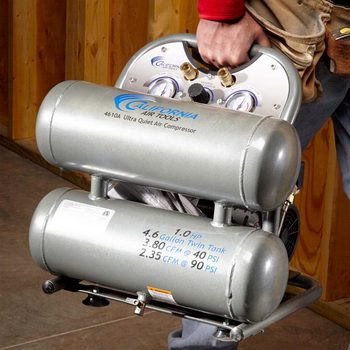 Air compressor being carried indoors; features dual tanks with specifications printed on them for performance details.