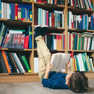 A child lies on the floor reading, with legs raised against a library bookshelf filled with colorful books.