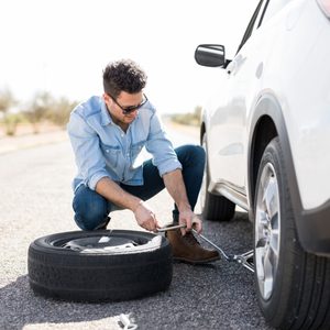Man crouched, changing a tire using a wrench beside a car on a deserted road, with sparse vegetation in the background.