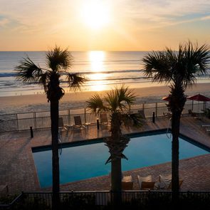 Swimming pool reflecting sunlight near palm trees, facing an ocean during a bright sunset. Beach chairs and umbrellas are scattered on a tiled patio.