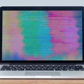 Laptop displays colorful glitching screen, resting on a wooden surface against a plain background.