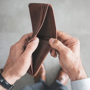 Hands hold an empty brown wallet, suggesting financial scarcity, against a neutral tiled floor background.