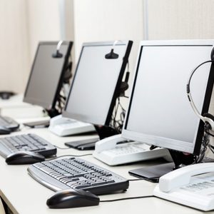 Desks display computer monitors with headsets and keyboards, alongside telephones, in a call center environment.