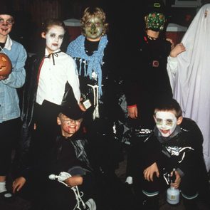 Children in Halloween costumes stand and crouch, posing together with face paint and masks in a dark outdoor setting. One holds a pumpkin.