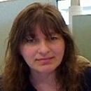 A person is sitting indoors, facing the camera, with long hair and a subtle smile. The background shows part of an office interior.