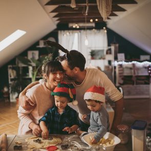 Family baking together, smiling, in a festive, decorated room with Christmas tree.