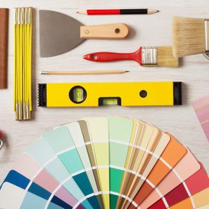 Various painting tools and materials lie arranged on a wooden surface; includes brushes, color swatches, a level, and a paint roller.