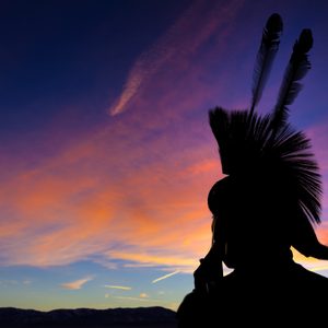 Silhouette of a person with feathered headdress stands against colorful sunset sky, with mountains in the background.
