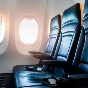 Airplane seats are lined up, empty, with seatbelts fastened on blue upholstery. Sunlight filters through two oval windows, illuminating the cabin interior.