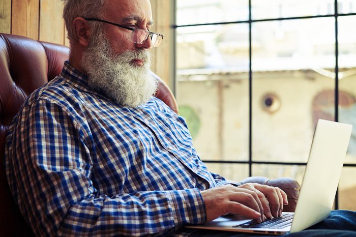 bearded senior man in glasses sitting on the couch and working with laptop at home