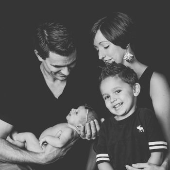 Parents and two children smile, with the father holding a newborn, in a black-and-white studio portrait.