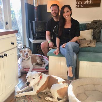 Couple sits smiling in cozy living room with two large dogs resting nearby; a wood stove and "i'm with you" sign in background.