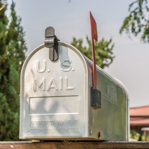 A silver mailbox stands with a raised flag, surrounded by greenery, under a clear sky. Text reads 